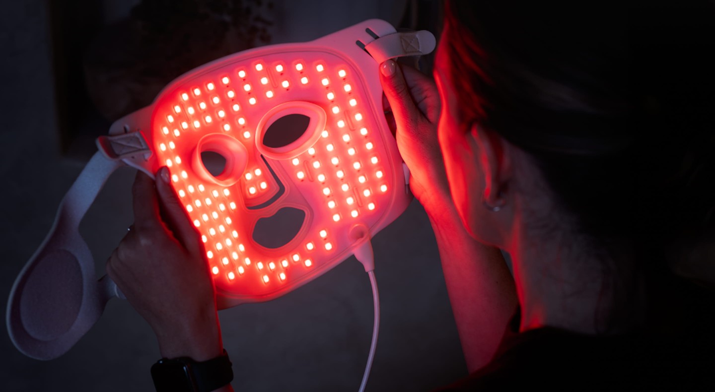 Person holding an LED face mask emitting bright red light, viewed from behind and slightly above their shoulder.