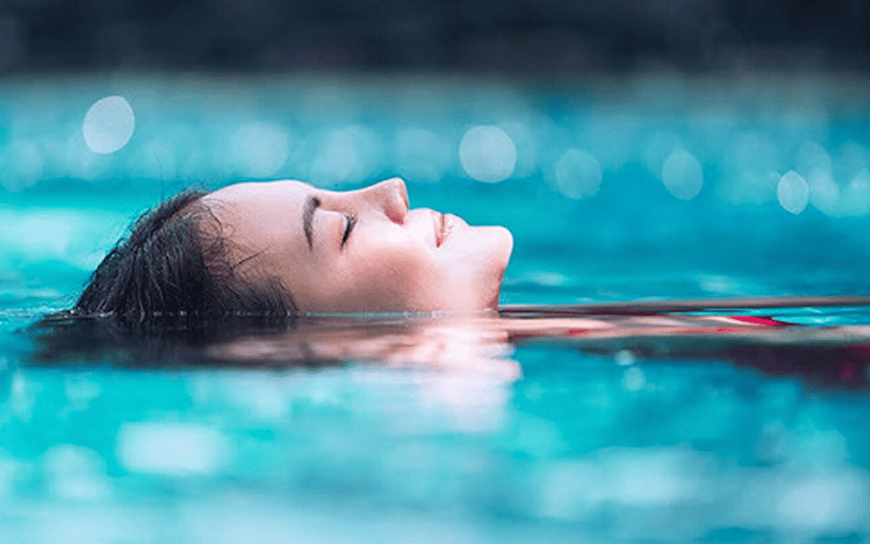 A woman floats on her back in a swimming pool with her eyes closed, appearing relaxed.