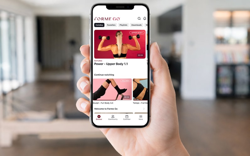 A person holds a smartphone displaying a fitness app with workout videos in a modern, bright living room.
